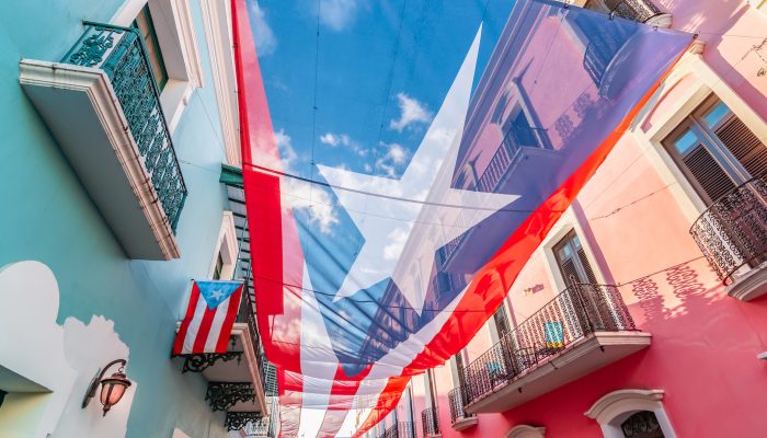 Colorful image of city centre of San Juan with large Puerto Rican flag above the street. Blue and pink buildings in the street. Sunny day. Red and white stripes, white star and blue colored national flag of Puerto Rico.